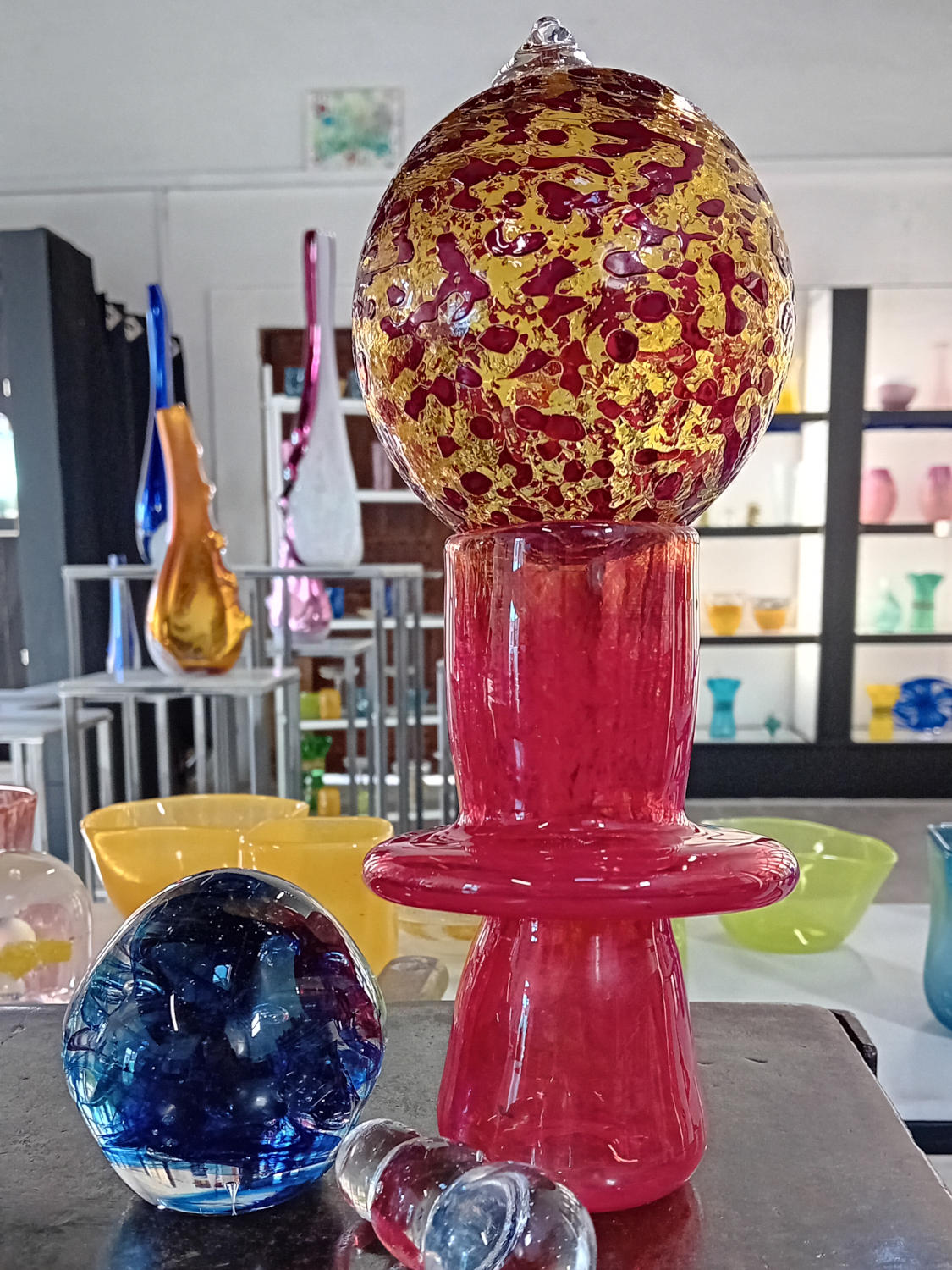 Colourful glass art pieces exhibited in a close-up in a workshop: a large blown glass sculpture composed of a red and purple base topped by a transparent sphere with pink reflections, accompanied by other decorative objects.
