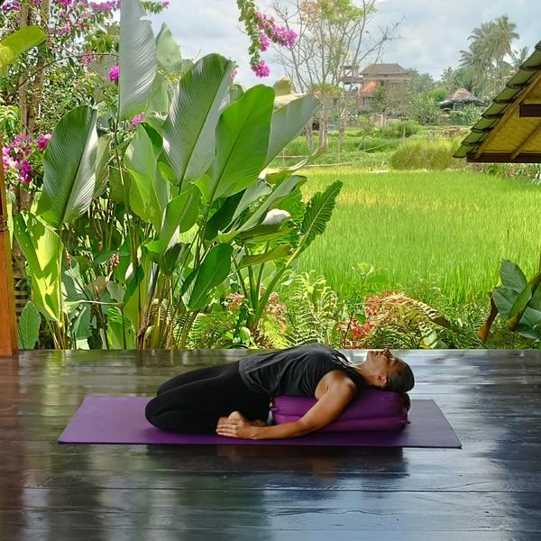 A person practices a restorative yoga pose, lying on a purple yoga mat with a bolster under their head and upper back, arms at their sides, in a verdant setting with lush vegetation and a field in the background.