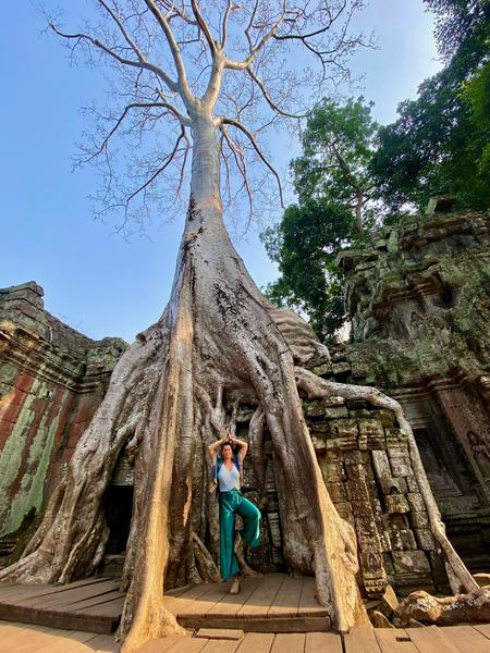 A woman poses in front of the massive roots of a large tree surrounding the ruins of Ta Prohm temple.