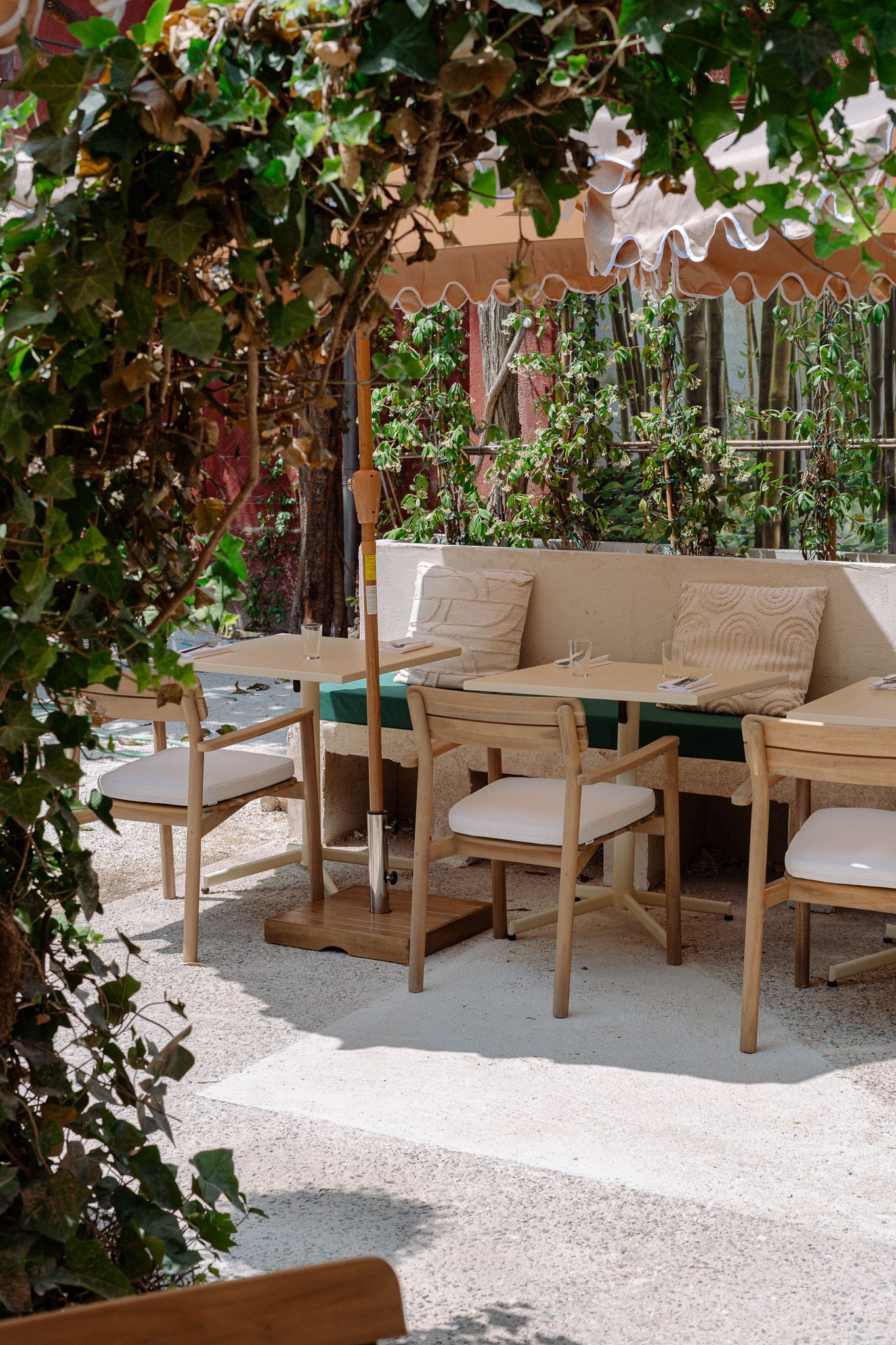 Café terrace shaded by greenery and an umbrella, with light wooden tables and upholstered seats.