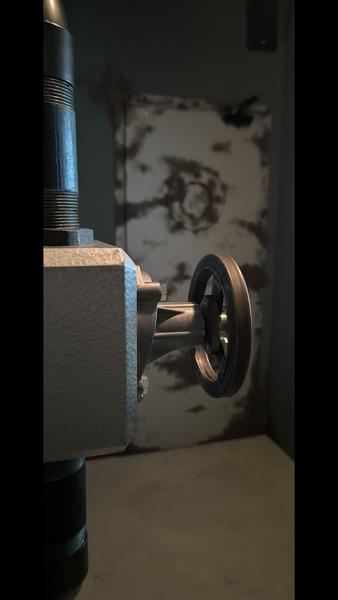 A close-up of a metal valve wheel in an industrial environment, with a rusty, textured door visible in the background.