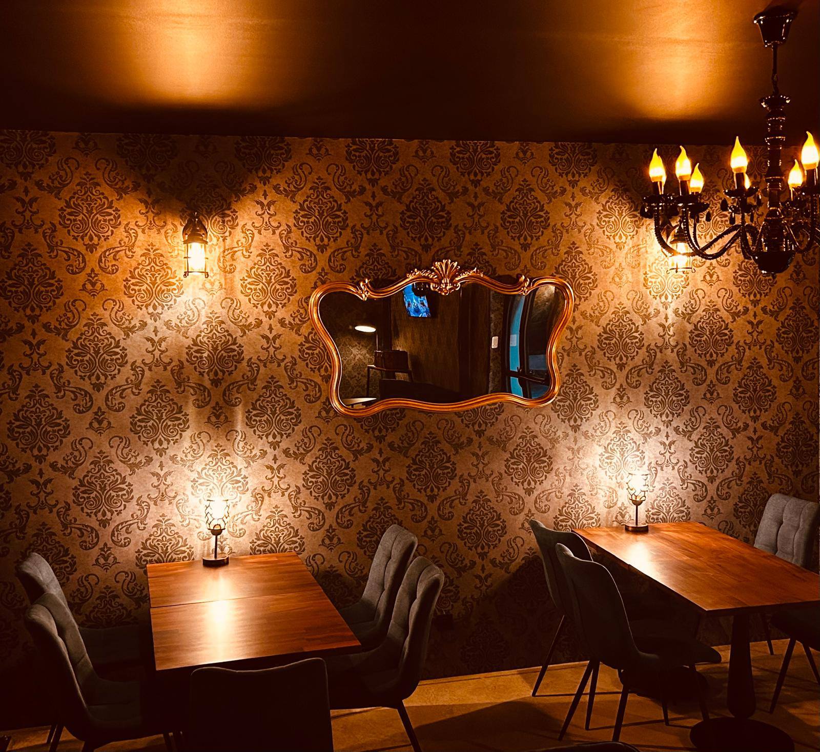 A restaurant or bar with Victorian decor, including tables and chairs, brown and gold damask wallpaper, a Rococo mirror on the wall, and a large chandelier on the ceiling.