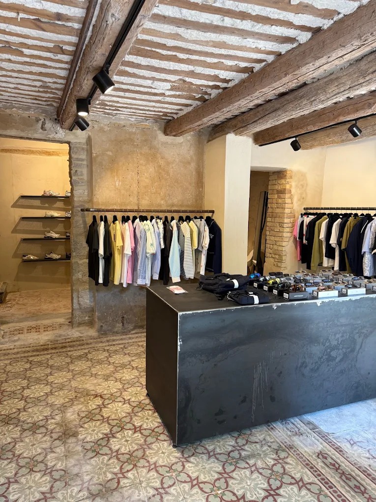 shop interior with ceiling beams, tiled floors, worn cement tiles, worn cement tiles, clothes racks and black metal central island with accessories on top: caps and glasses