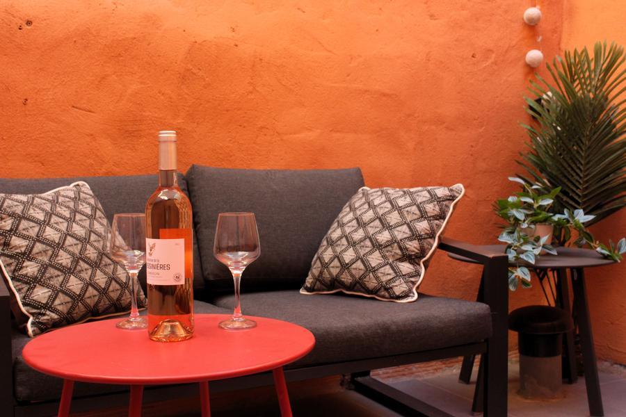 A small patio with orange walls, warm and intimate. A dark gray sofa with two black and white geometric cushions occupies the background of the scene. In front, a round red coffee table holds a bottle of rosé wine and two stemmed glasses.. On the right, a side table accommodates a green potted plant, adding a touch of freshness. The atmosphere evokes a moment of convivial relaxation, perfect for an aperitif. 