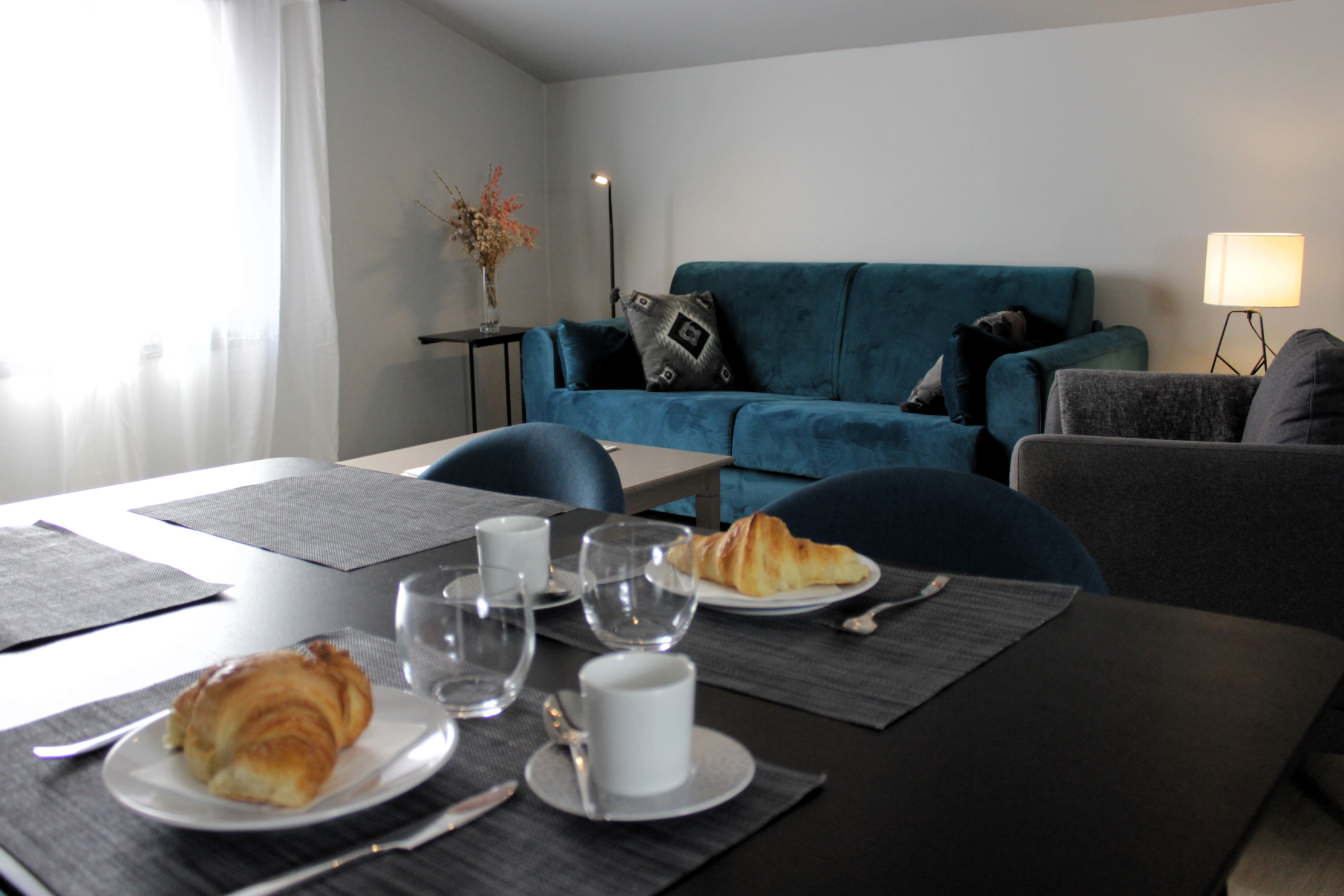 A bright, modern room decorated in a simple and elegant style. In the foreground, a black breakfast table is set up. In the foreground, a black breakfast table with two plates containing croissants, round glasses and white cups on gray placemats. In the background, a turquoise velvet sofa is decorated with black and gray geometric cushions. On the right, a table lamp diffuses soft light, and a bouquet of dried flowers decorates a coffee table. The set creates a welcoming and cozy atmosphere. 