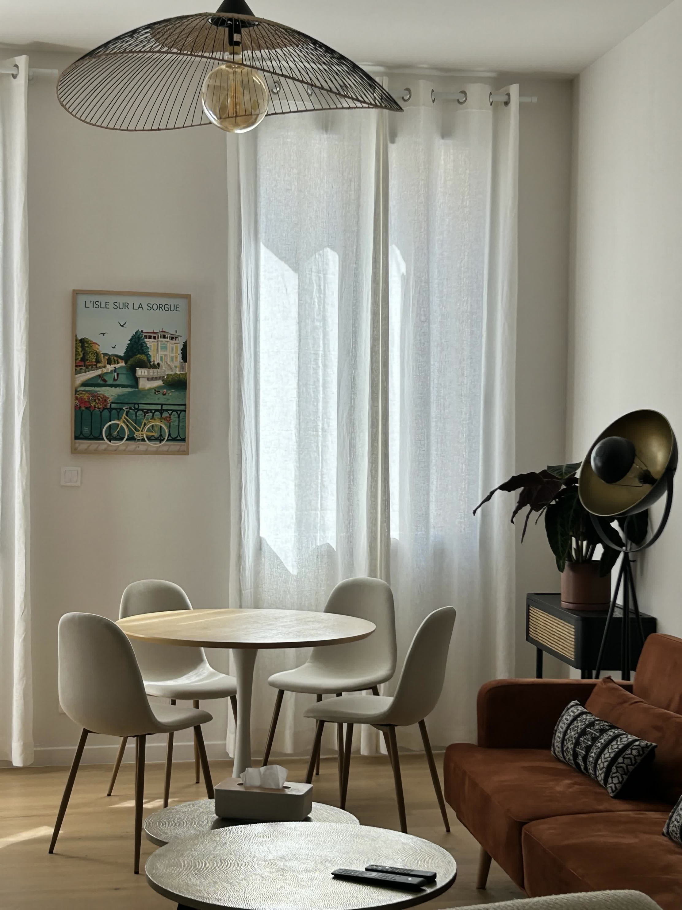 Bright living room with a dining area composed of a round table and light chairs, a brown sofa, a large veiled window and modern decor in natural tones.
