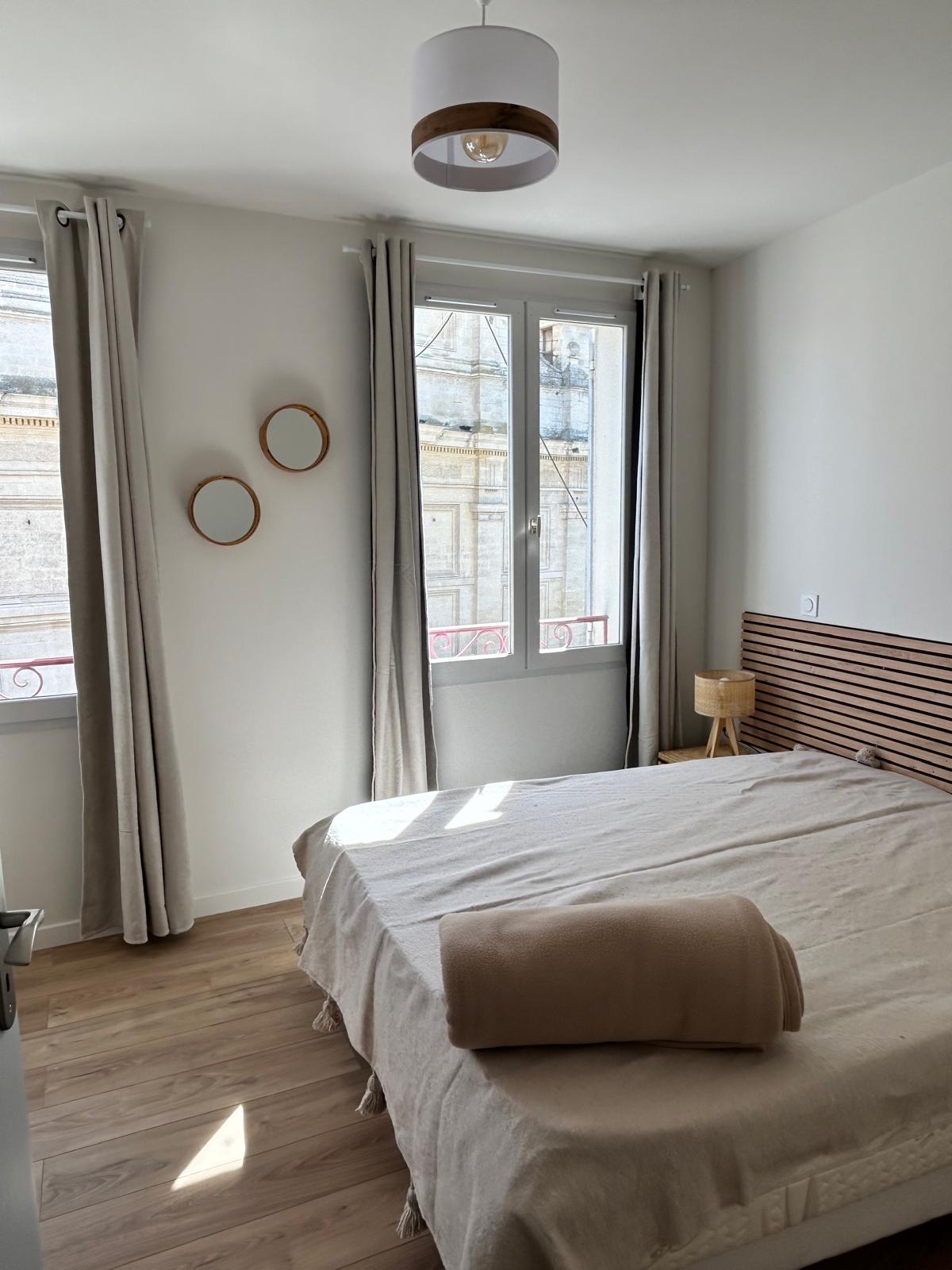 Sleek and bright bedroom with a double bed in beige textiles, a wooden headboard, light curtains and a large window that lets in daylight.