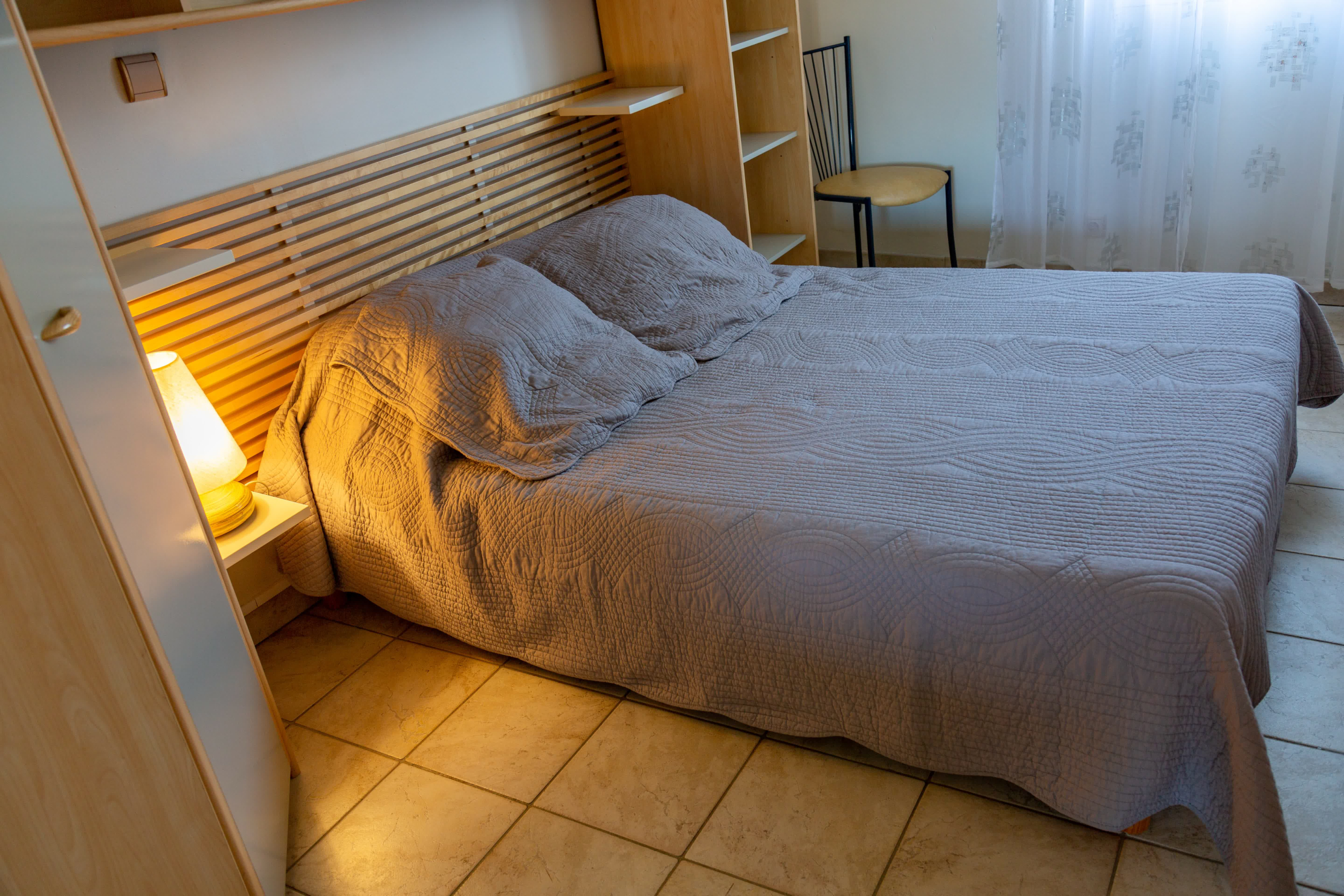Quiet room with a double bed in neutral tones, a light wooden headboard, built-in storage, and soft light from a lamp and a veiled window.