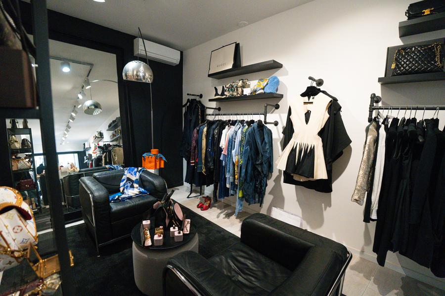 The photo shows the interior of a high-end clothing store with a contemporary and refined style. In the foreground, there is a cozy space composed of black leather armchairs arranged around a small coffee table, creating an almost private atmosphere, like a living room. On the left, bags from major brands are displayed, suggesting a luxury or premium second-hand offer. In the background and on the walls, several racks present stylish clothing. neatly arranged, mostly in sober tones (black, white, denim). A structured two-tone dress attracts particular attention, highlighted as a key piece. Wall shelves also accommodate designer accessories and bags. 