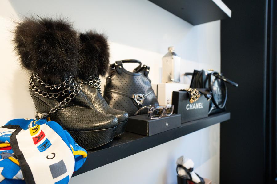 The photo shows an exhibition shelf showcasing a selection of high-end fashion accessories in an elegant and refined atmosphere. In the foreground, a pair of black boots catches the eye immediately. They stand out for their elaborate texture, their decorative metal chains and finished with fur, adding a touch that is both daring and sophisticated. Nearby, we see a colorful scarf that contrasts and with the dominant dark tones. On the shelf, several black leather handbags are carefully arranged, accompanied by accessories such as glasses and small leather goods. A Chanel box is also visible, reinforcing the image of a selection from major luxury houses. The set is organized in a minimalist manner, with a careful layout that highlights each piece. The general atmosphere evokes a chic and contemporary universe, typical of a store specializing in designer accessories or consignment shops high-end. 