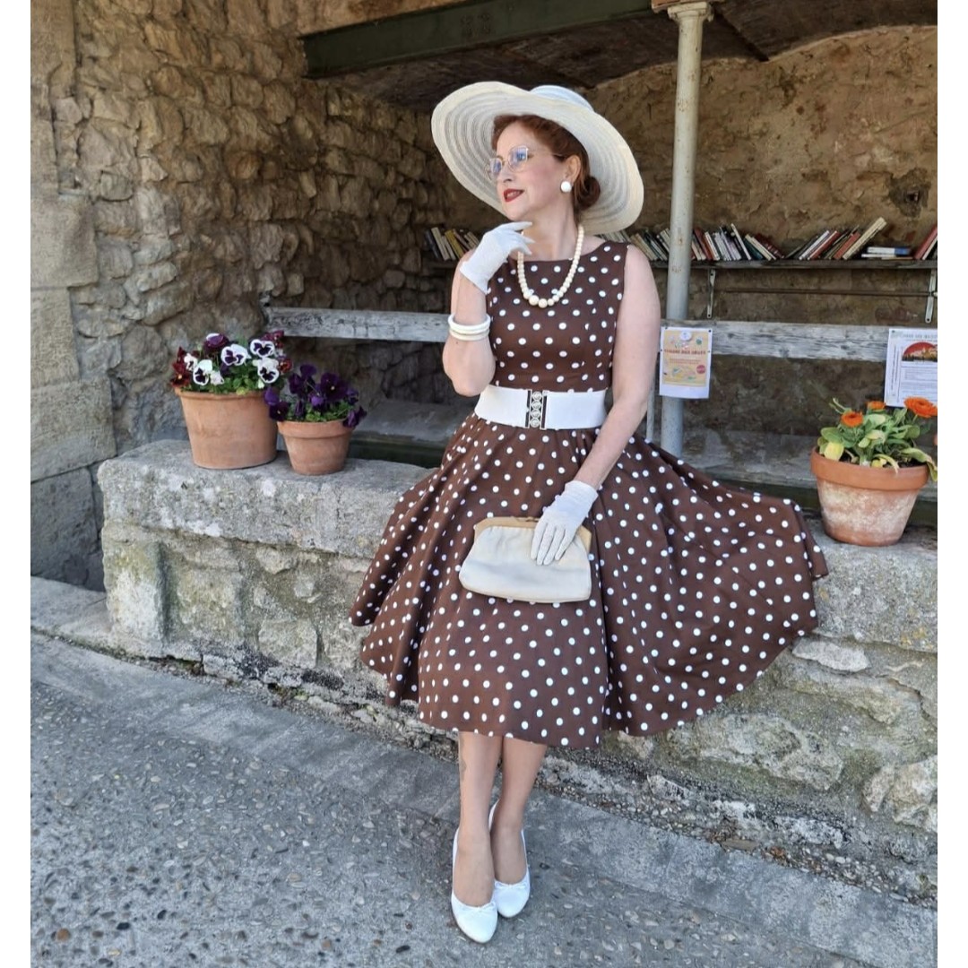woman sitting on a stone wall wearing a brown white polka dot dress with a brown white polka dot dress with hat, wide belt, hat, wide belt, white bag, gloves and pumps