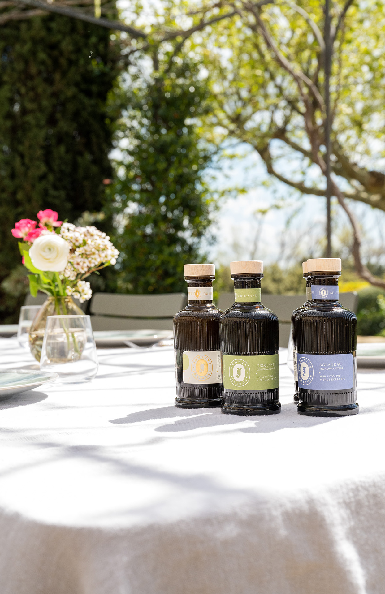 Three bottles of olive oil are placed on a large table covered with a white tablecloth, in a bright garden. Next to it, a small vase contains some fresh flowers. In the background, you can see trees and a sunny terrace. The bottles are elegant, dark, with colorful and refined labels. The set gives the impression of outdoor dining, careful eating and the Mediterranean lifestyle.