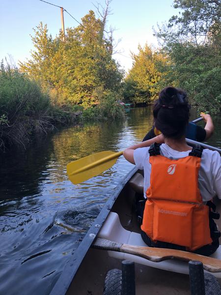 A person sitting in the back of a canoe advances gently on a calm river surrounded by trees and tall grass. The golden light at the end of the day is reflected on the water, creating a peaceful and immersive atmosphere in the heart of nature.