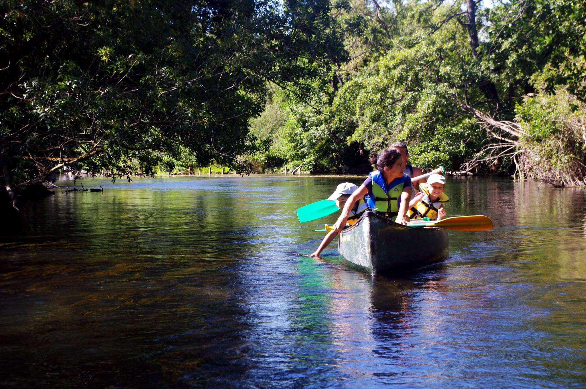 A family sails in canoes on a river with transparent waters bordered by dense and green vegetation. The adults paddle while the children enjoy the ride in a cheerful and refreshing atmosphere.