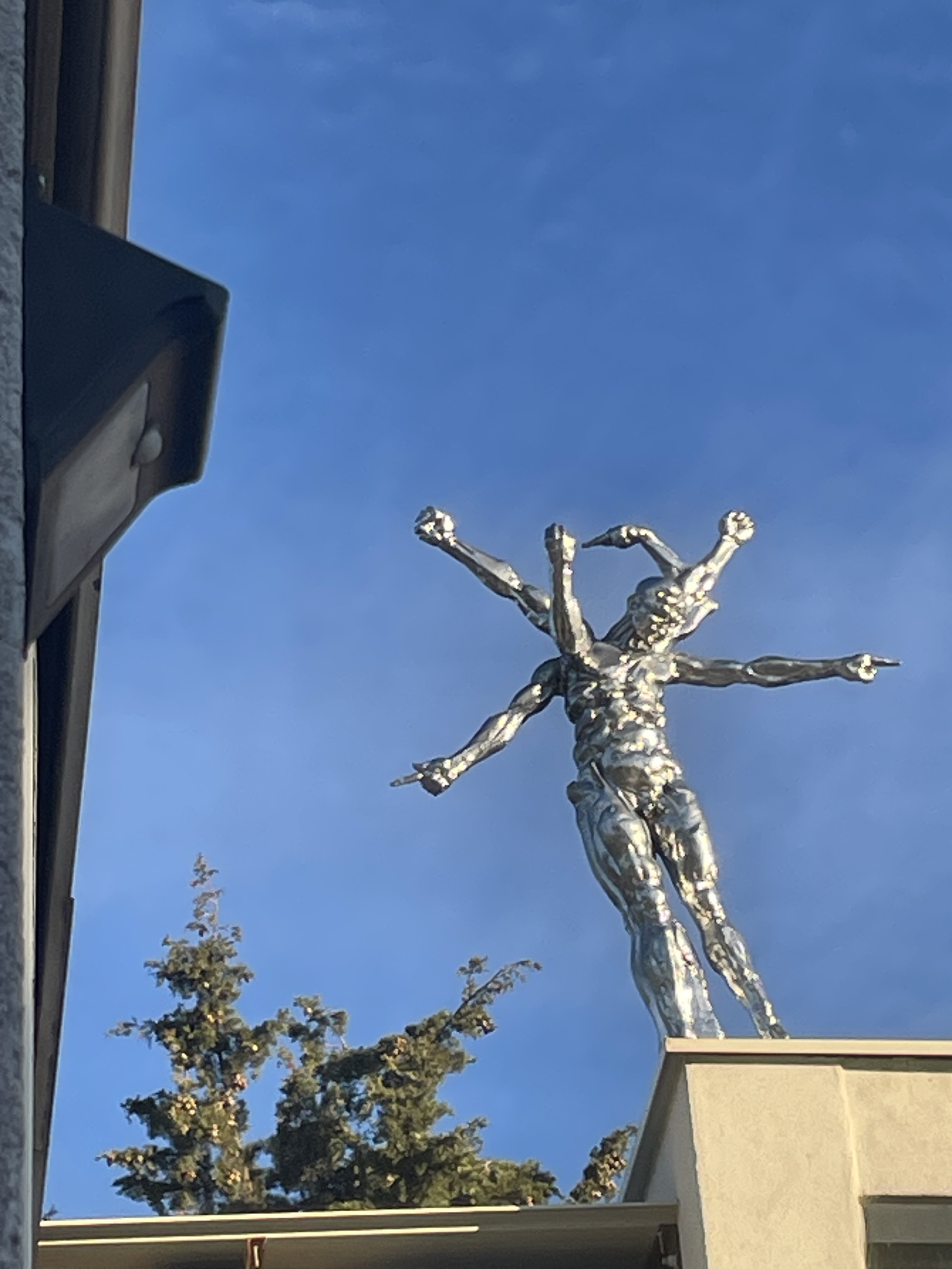 This photograph highlights a monumental sculpture representing a human figure with multiple arms, made in a shiny metallic finish that catches the light. Installed high up on a building, the
