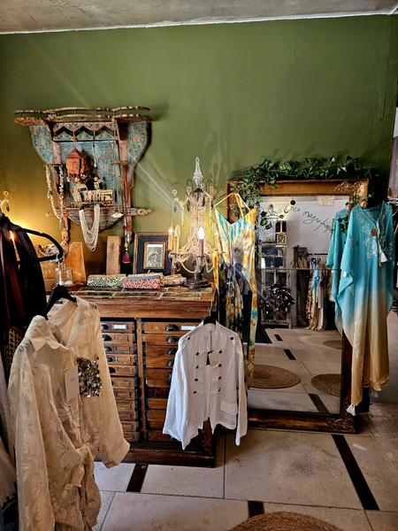 This space features several garments with bohemian inspirations: light shirts, flowing pieces, and brightly colored outfits. The antique furniture, the gilded mirror and the decorative objects create a singular decor that is both elegant and eclectic.