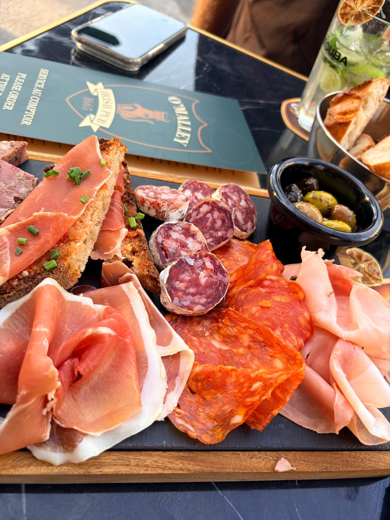 A generous charcuterie board to share, composed of hams, sausages and other specialties, accompanied by olives and bread. In the background, a cocktail and the O