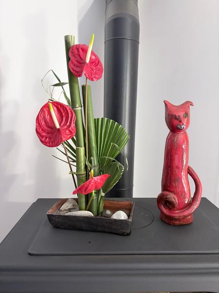Ikebana-style floral arrangement combining red anthuriums, bamboo stems and graphic foliage, arranged in a rectangular container with pebbles.
